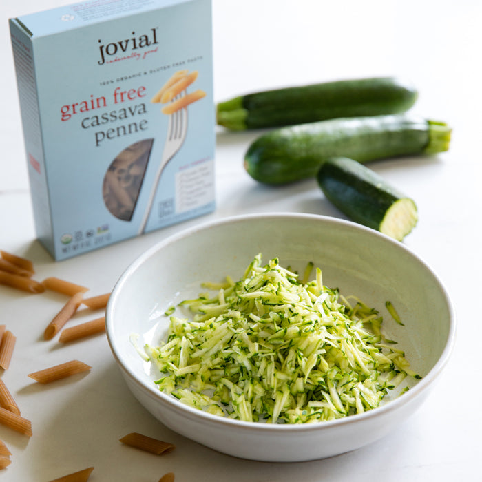 Bowl of shredded zucchini with a Jovial grain-free cassava penne box, whole zucchinis and raw penne on a white counter