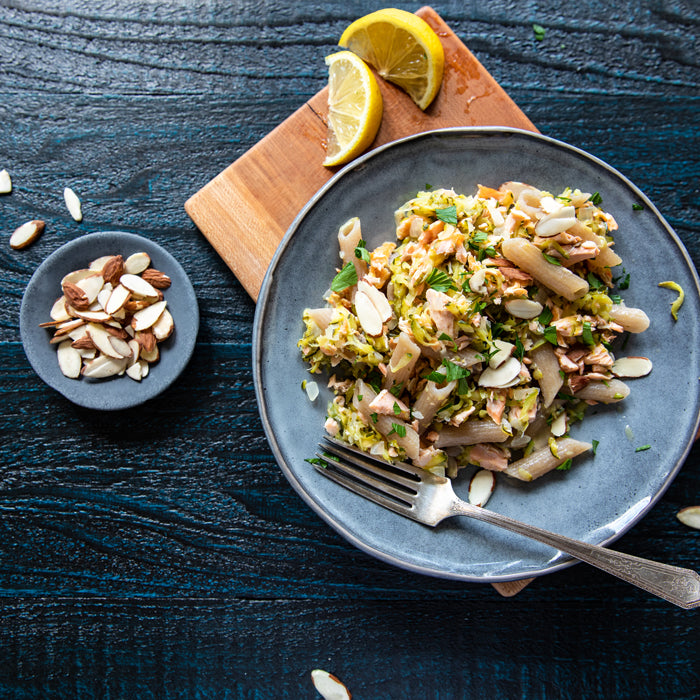 Salmon and zucchini cassava pasta topped with flaked salmon, sliced almonds, and parsley on a blue plate with lemon wedges