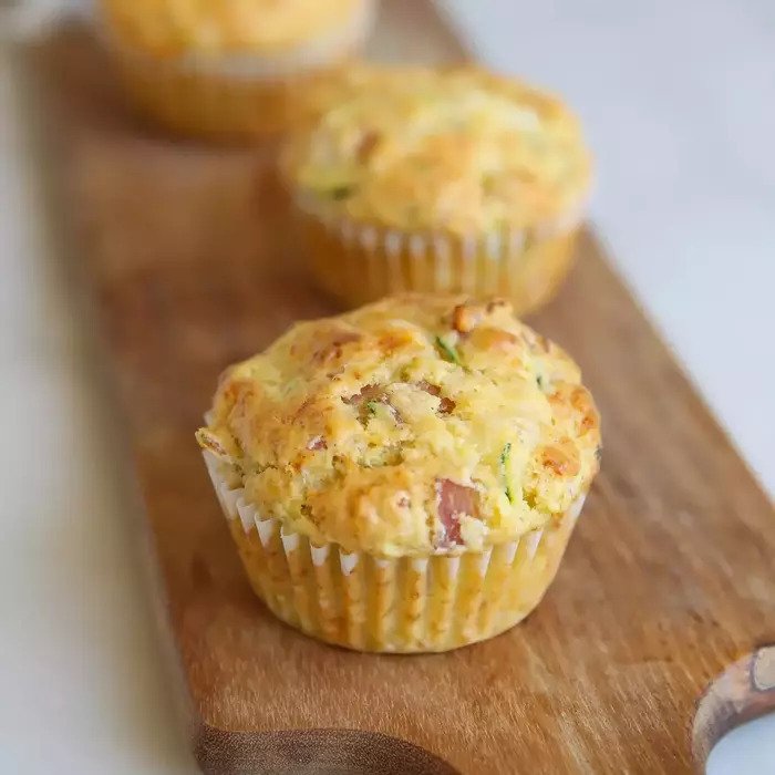 Close-up front view of a ham and cheese einkorn zucchini muffin showing golden top with visible zucchini and ham pieces on a wooden board