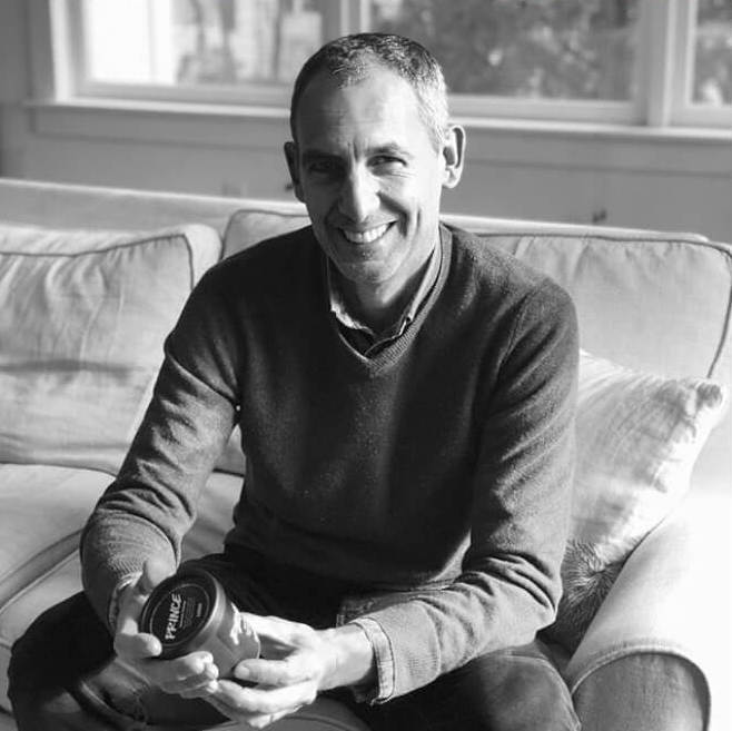 Man sitting on a sofa smiling while holding a jar of Jovial product in a sunlit living room