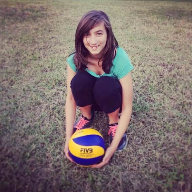 Young woman crouching on grass holding a yellow and blue volleyball and smiling at the camera