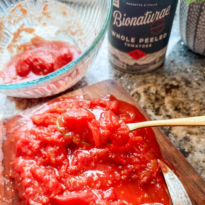 Spoon mashing canned whole tomatoes on a cutting board with a glass mixing bowl and can on a granite kitchen countertop