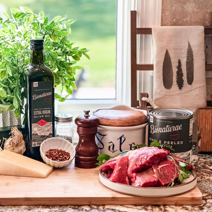 Raw steak and ingredients for pizzaiola on a cutting board with Bionaturae olive oil and tomatoes by a sunlit kitchen window