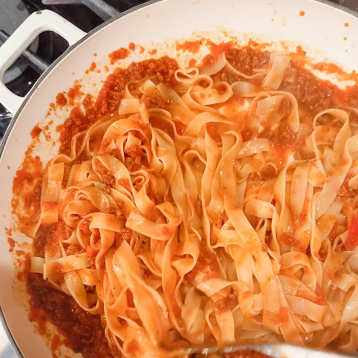 Tagliatelle tossed with Tuscan ragù in a white enameled skillet, thick tomato and meat sauce coating the noodles
