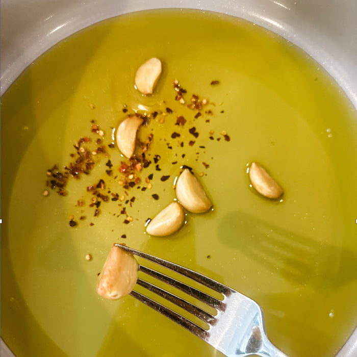Garlic cloves in olive oil with red pepper flakes in a white bowl, one clove held on a fork