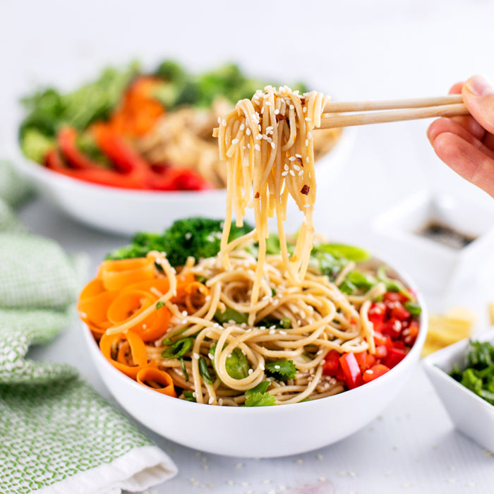 Chopsticks lifting sesame scallion ginger noodles from a white bowl topped with carrot ribbons, broccoli, and red pepper