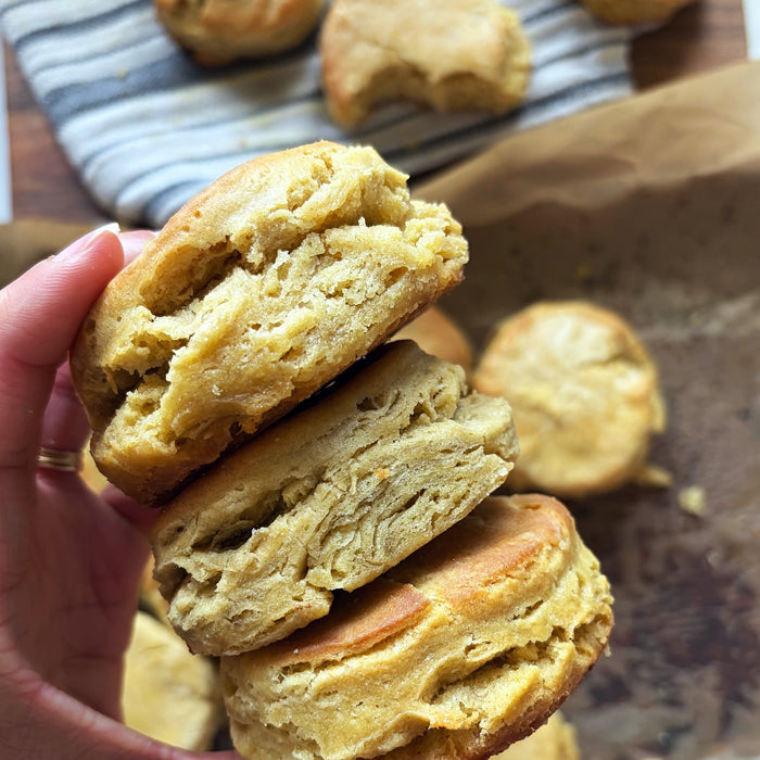 Close-up of stacked einkorn sourdough biscuits showing flaky layers and golden crust, hand holding with parchment and striped cloth background