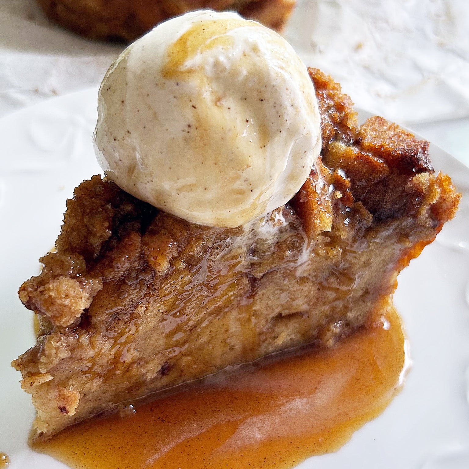 Close-up side view of maple caramel sourdough bread pudding topped with vanilla ice cream and caramel sauce on a white plate