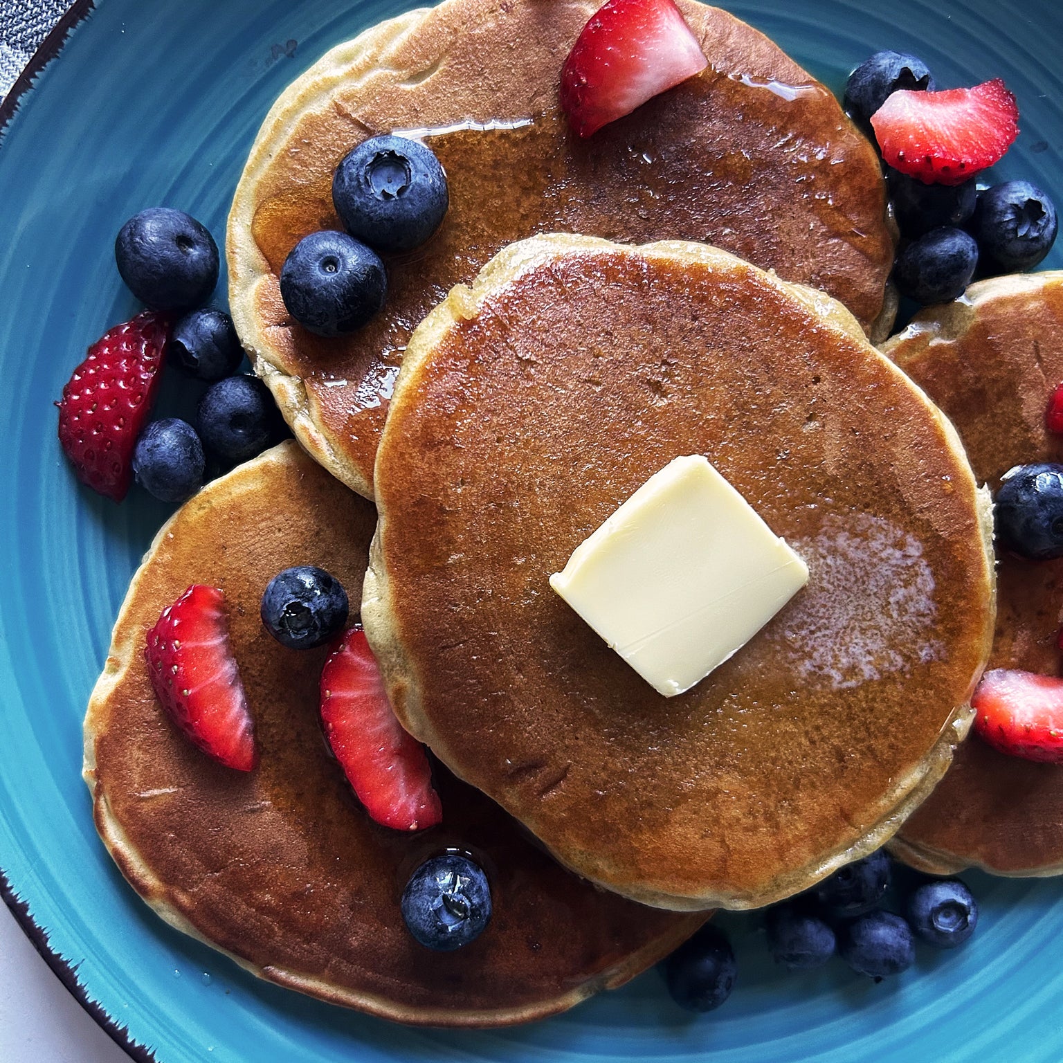 Top-down view of sourdough Einkorn pancakes with butter and syrup, sliced strawberries and blueberries on a blue plate
