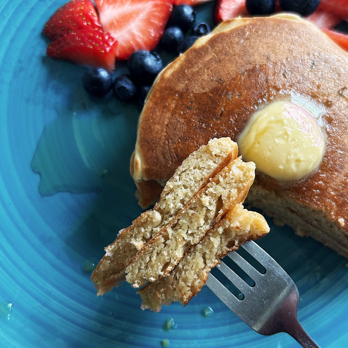 Fork holding a bite of sourdough Einkorn pancakes with butter, maple syrup, and berries on a turquoise plate