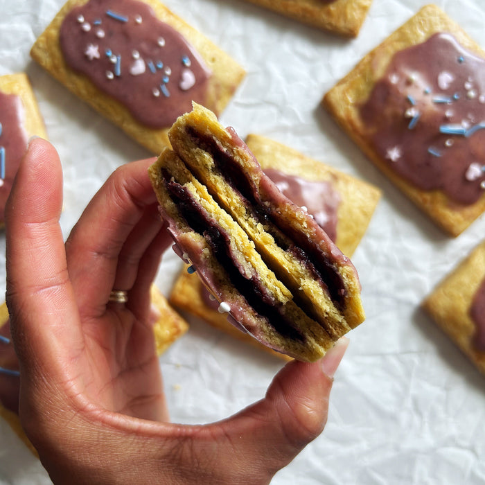 Close-up of hand holding a torn-open sourdough wild berry pop tart showing jam filling and purple icing with sprinkles on parchment