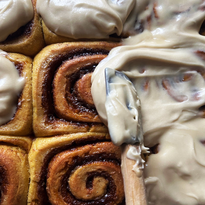 Close-up overhead of sourdough pumpkin rolls showing cinnamon swirls and a spatula spreading maple cream cheese frosting