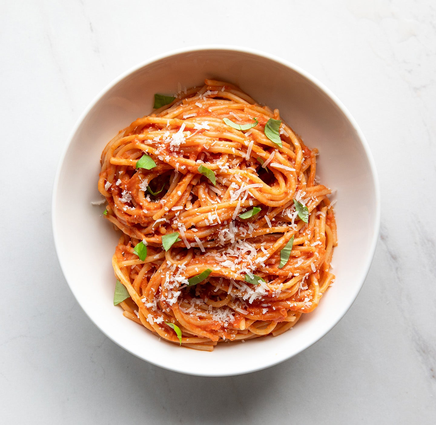 Bowl of sourdough spaghetti tossed in tomato sauce, garnished with grated cheese and basil on a white marble surface