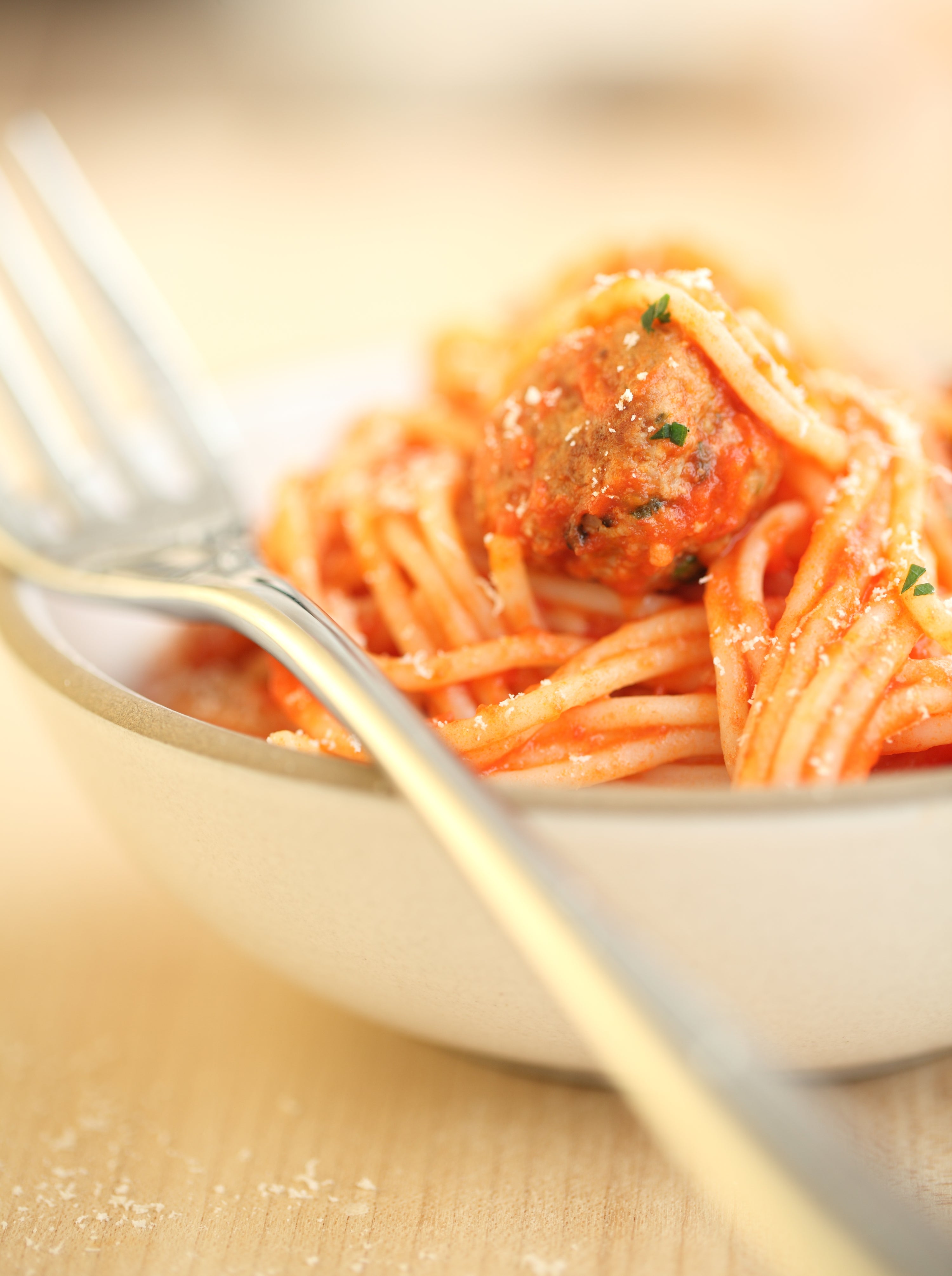 Bowl of gluten-free spaghetti twirled on a fork, topped with a meatball and grated cheese on a wooden table
