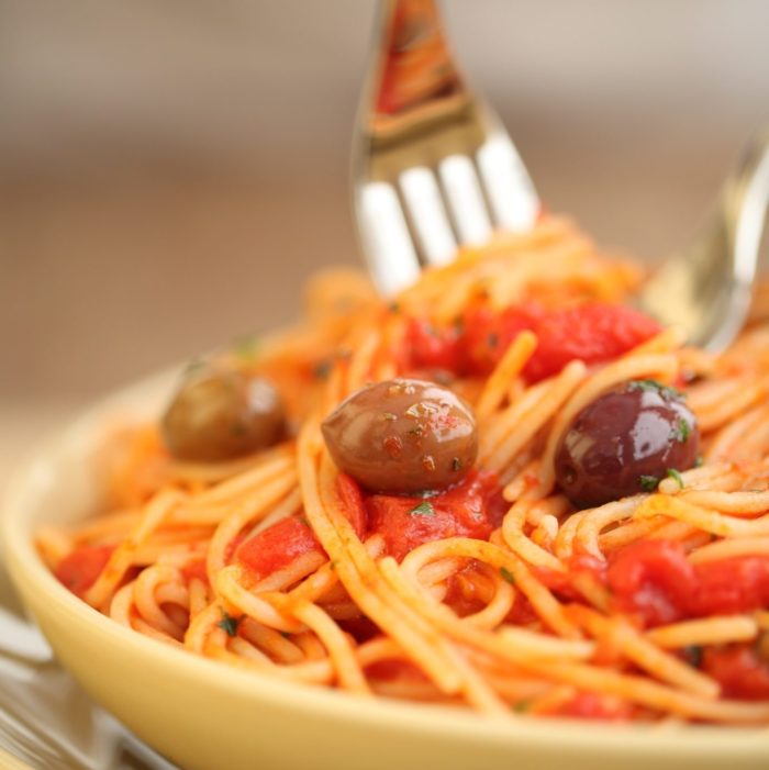 Brown rice spaghetti puttanesca twirled on a fork over a shallow bowl with tomatoes, basil, and olives