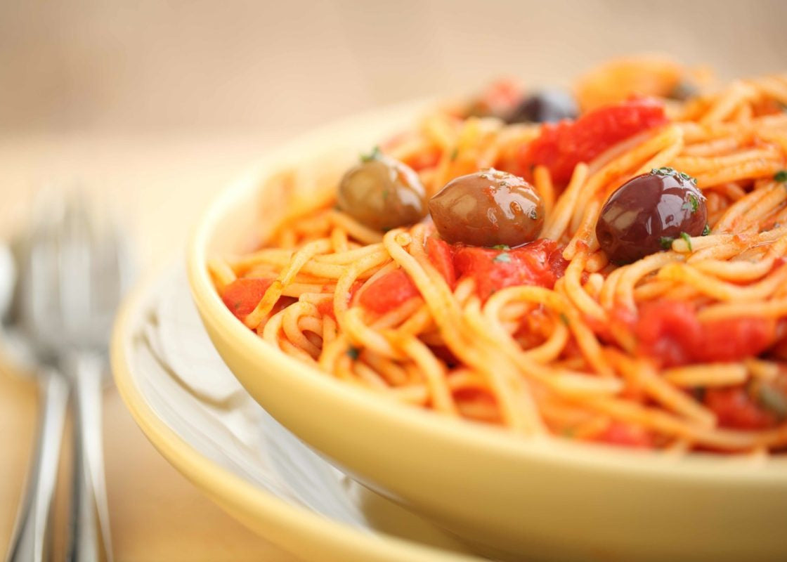 Spaghetti puttanesca with tomatoes, olives, and basil served in a yellow bowl on a wooden table with a fork in the background