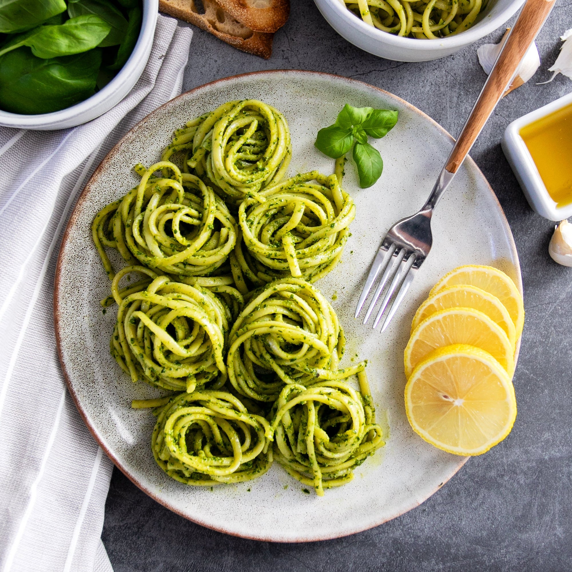Top-down view of Organic Gluten Free Rice & Lentil Linguine tossed in spinach pesto, plated as six nests with lemon slices and a fork on a gray plate