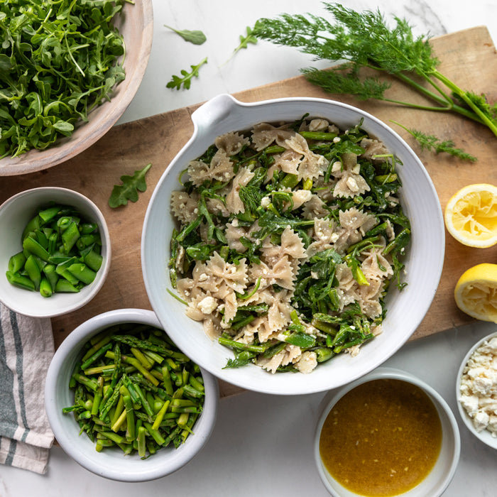 Spring pasta salad with gluten free brown rice bowtie pasta, asparagus, snap peas, arugula, and feta in a white bowl with lemon and dressing