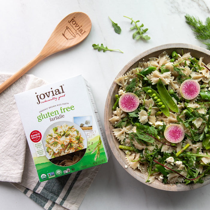 Top-down view of Jovial gluten free brown rice farfalle box beside a bowl of spring pasta salad showcasing asparagus, sugar snap peas, dill, feta, and radish on a marble surface