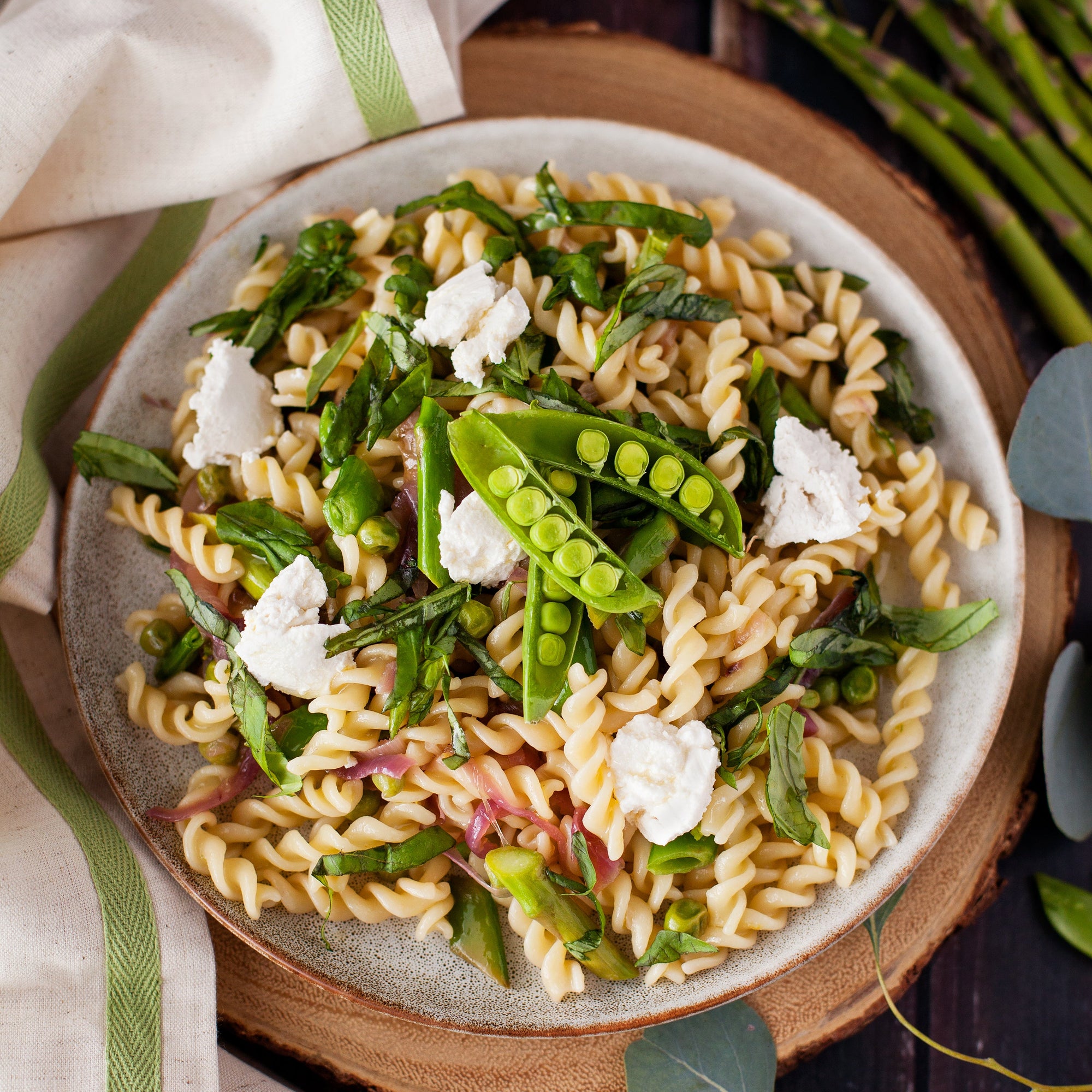 Bowl of cooked organic sourdough fusilli tossed with sugar snap peas, fresh greens, and dollops of ricotta on a wooden board