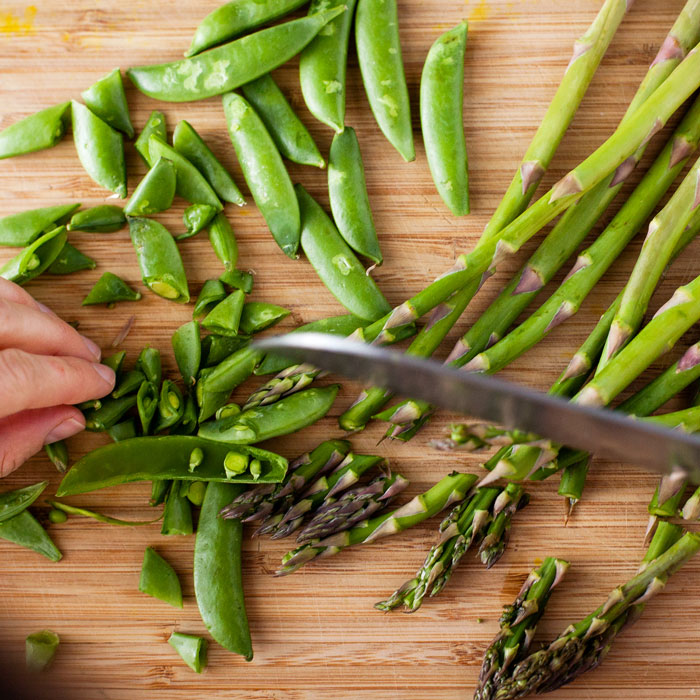 Hands slicing snap peas and trimming asparagus on a wooden cutting board for spring vegetable fusilli