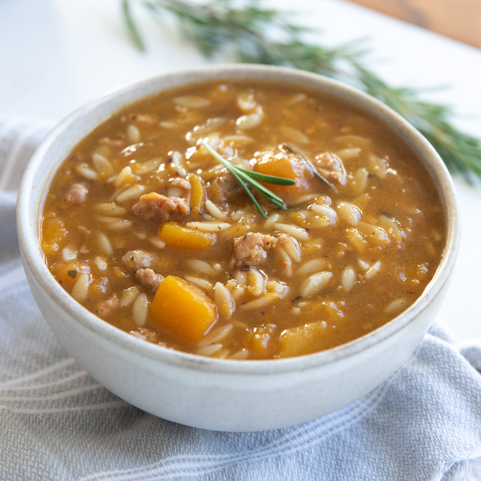 Angled close-up of autumn squash and orzo soup with sausage, with squash cubes, sausage, and a rosemary sprig in a white bowl