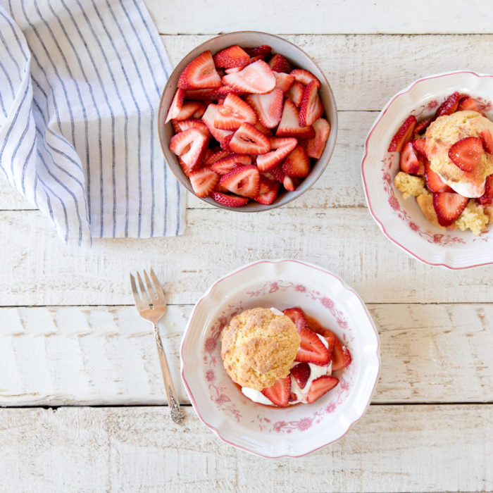 Einkorn strawberry shortcakes served with whipped cream and sliced strawberries in floral bowls on a white wooden table