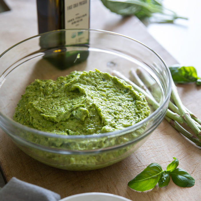 Close-up of a glass bowl of organic asparagus pesto showcasing its creamy texture with an olive oil bottle and asparagus on a wooden cutting board
