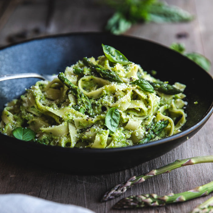 Egg tagliatelle tossed with asparagus pesto, asparagus tips, grated cheese, and basil in a black bowl on a wooden table