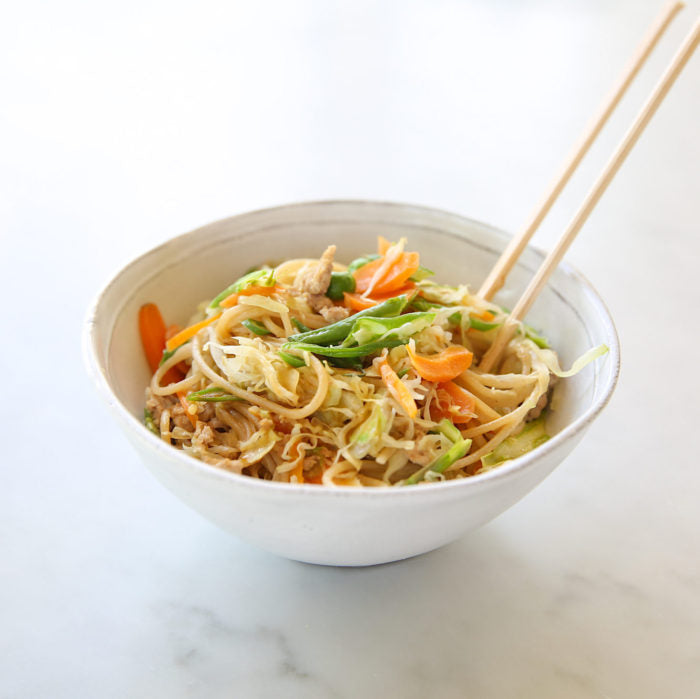 Top-front view of teriyaki brown rice pasta stir-fry in a white bowl with cabbage, carrots, snap peas and chopsticks