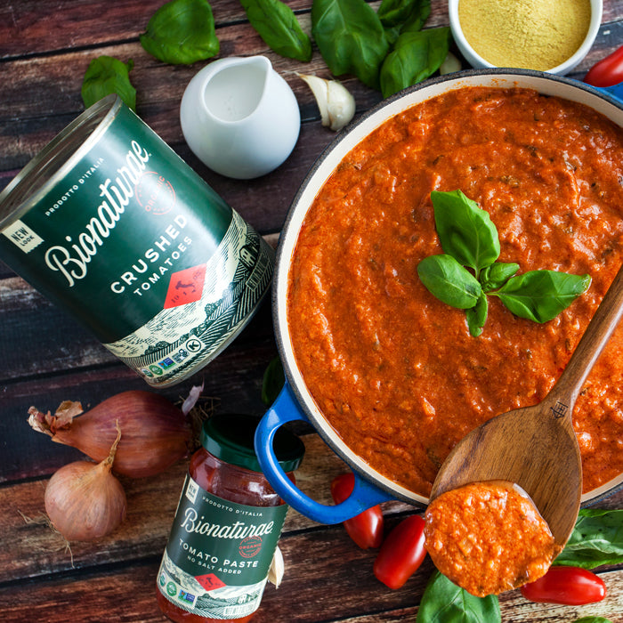 Tomato basil cream sauce in a blue skillet with wooden spoon, fresh basil, and Bionaturae crushed tomatoes and tomato paste