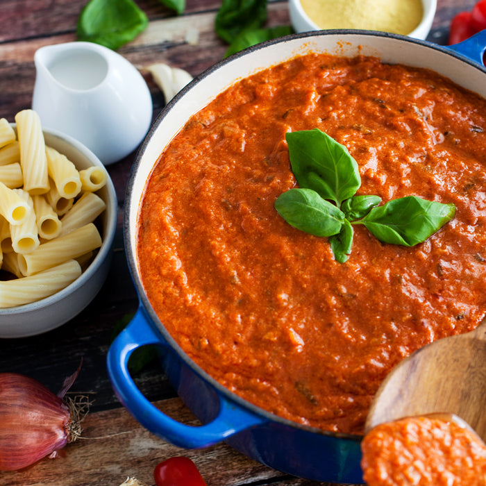 Top-down view of tomato basil cream sauce in a blue Dutch oven showcasing creamy texture and fresh basil garnish, with a bowl of rigatoni and a wooden spoon