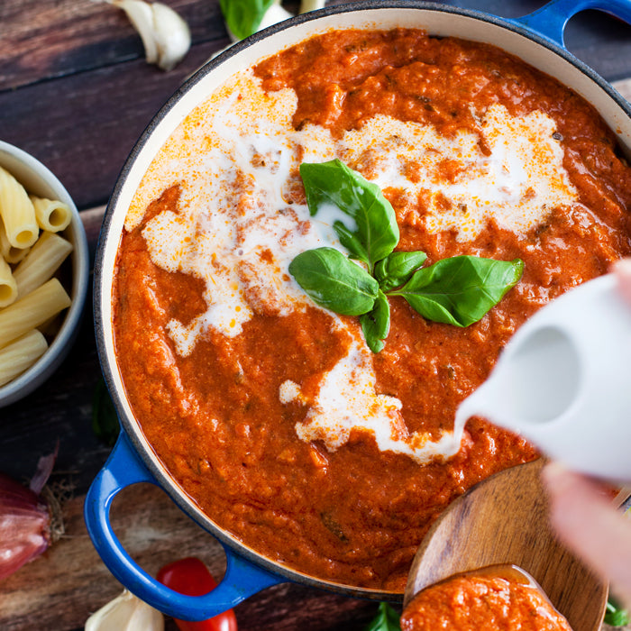 Top-down view of Italian tomato basil cream sauce in a blue pot as cream is poured, basil garnish, penne and garlic nearby