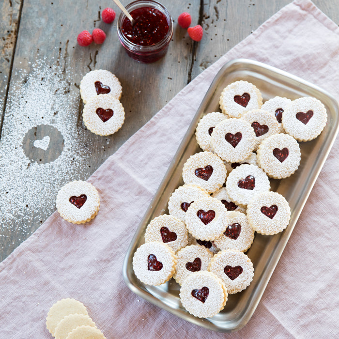 Overhead view of Einkorn Linzer cookies showcasing heart-shaped raspberry jam centers and powdered sugar on a metal tray