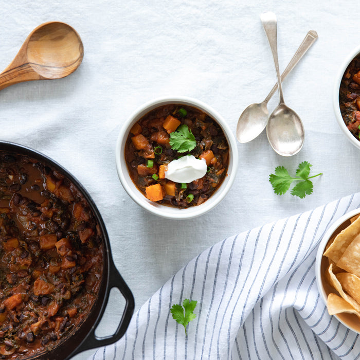 Bowl of organic vegetarian chili with butternut squash and kale, topped with sour cream and cilantro, next to a cast-iron pot