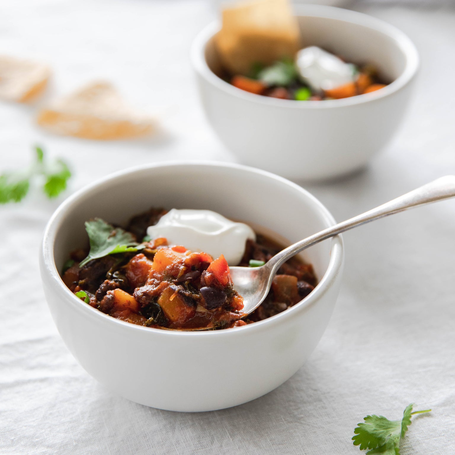Bowl of vegetarian chili with butternut squash and kale, topped with sour cream and cilantro, spoon in bowl on white cloth