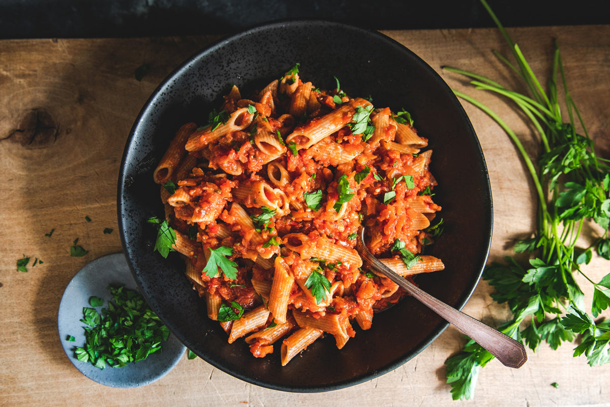 Bowl of penne with eggplant bolognese with a fork, served on a wooden table with fresh parsley garnish
