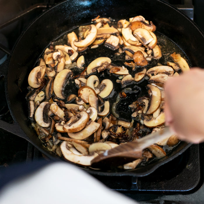 Sliced brown mushrooms sautéing in a cast-iron skillet on a stovetop, being stirred with a wooden spoon