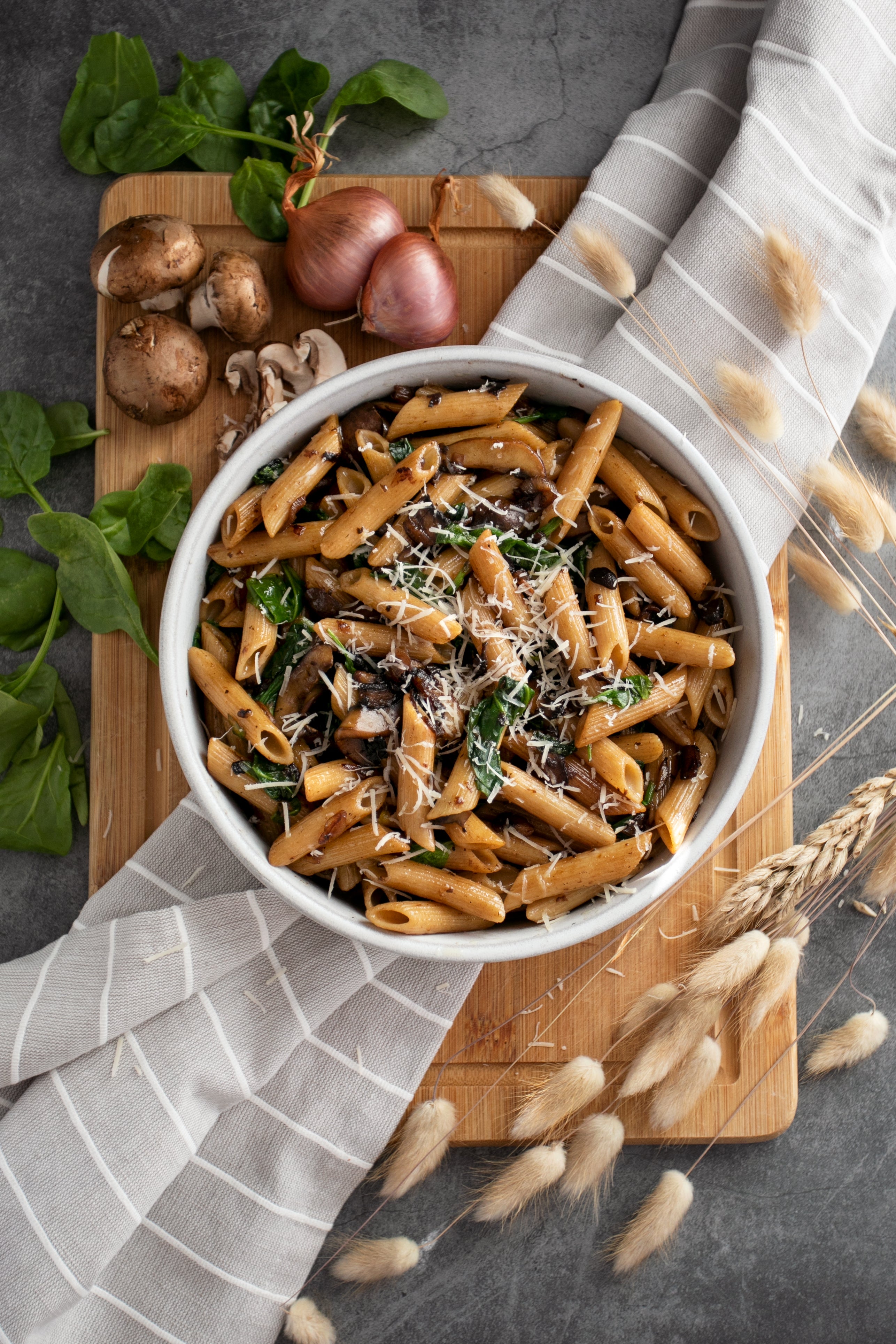 Top-down view of brown rice penne with sauteed mushrooms, spinach, and grated cheese in a bowl on a wooden board