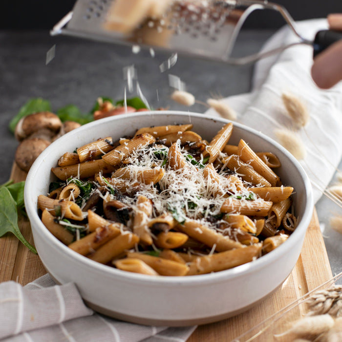 Whole wheat penne with sautéed mushrooms and wilted spinach in a bowl, grated cheese being sprinkled on top on a wooden board