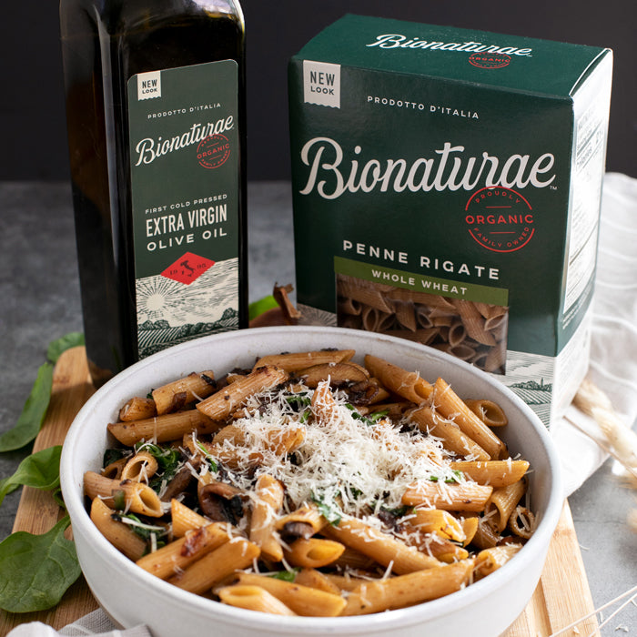 Bowl of whole wheat penne with sauteed mushrooms, garlicky spinach, and grated cheese beside a Bionaturae pasta box and olive oil bottle