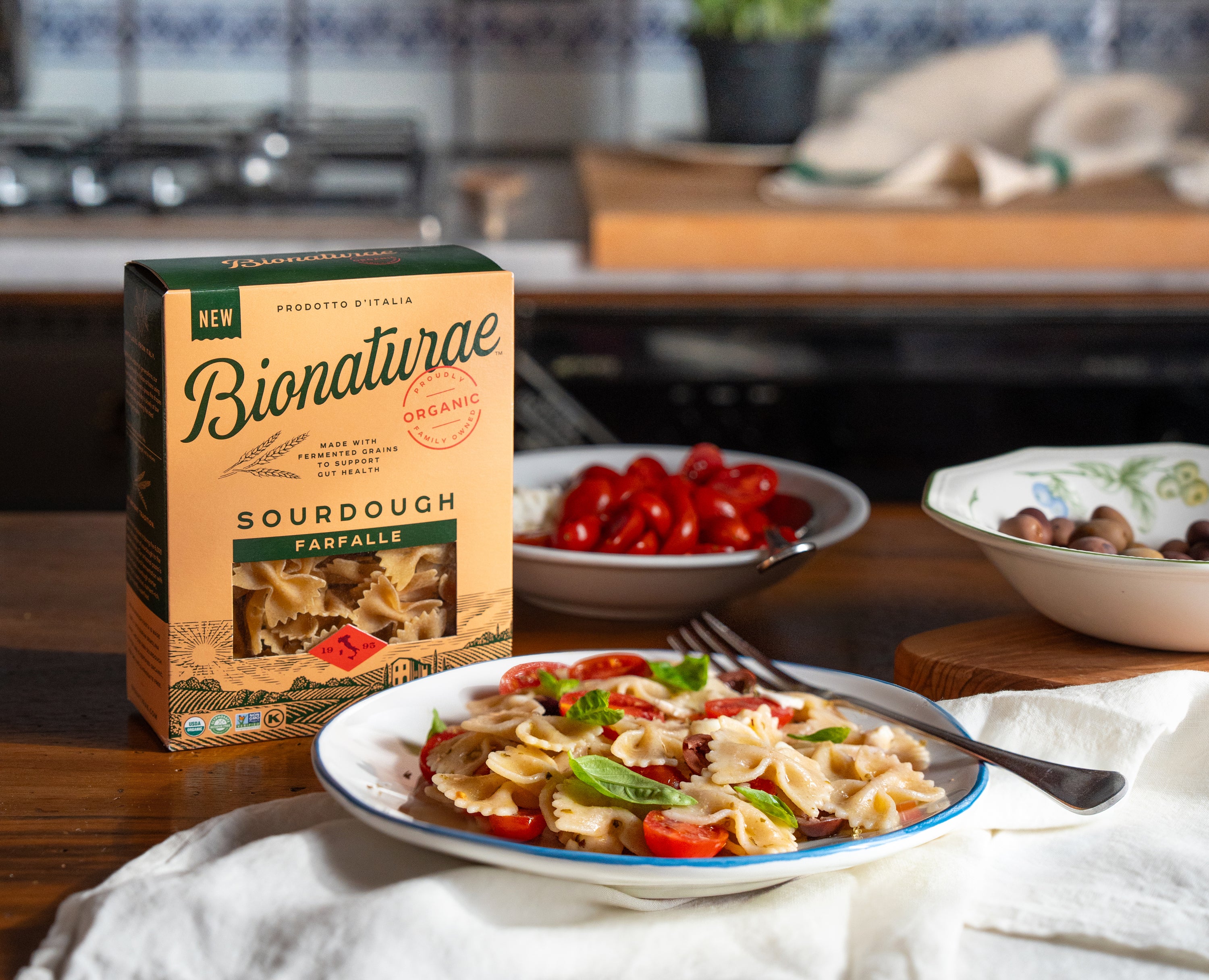 Front view of Organic Bionaturae farfalle box with window, plated farfalle with cherry tomatoes and basil on wooden counter