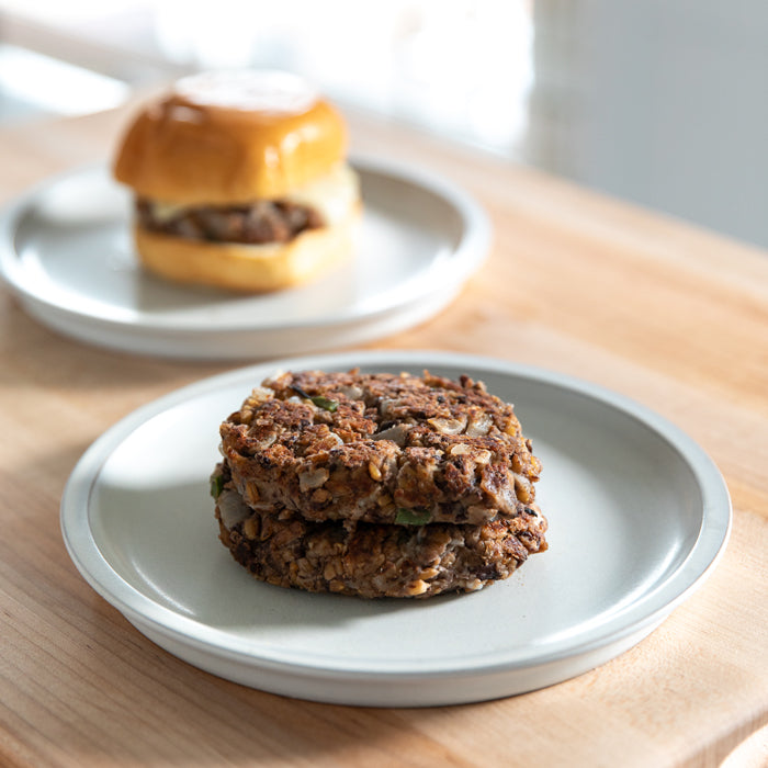 Two black-bean and Einkorn wheat-berry patties stacked on a ceramic plate with a burger on a bun blurred in the background
