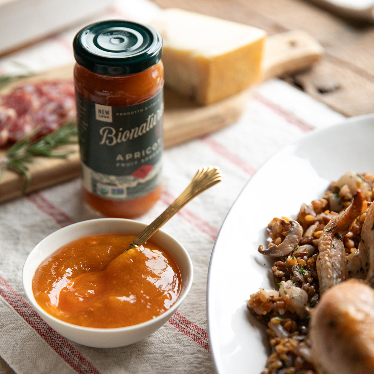 Jar of organic apricot fruit spread beside a bowl of spread with a spoon and a plate of wheat-berry stuffing on a rustic table