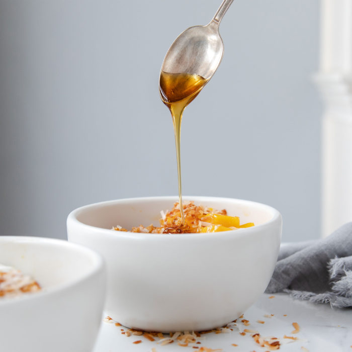 Wheat berries topped with diced mango and toasted coconut, honey drizzling from a spoon into a white bowl on a light surface