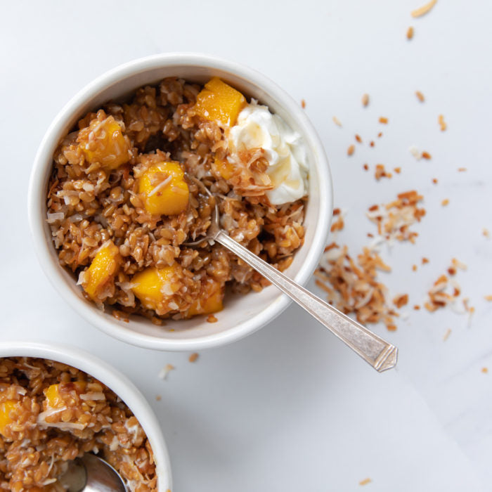 Top-down view of wheat berry breakfast bowl with mango, toasted coconut, yogurt, and spoon on white background