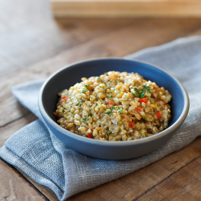 Cooked wheat berries in a blue-gray bowl on a wooden table, garnished with chopped herbs and red pepper on a folded gray napkin