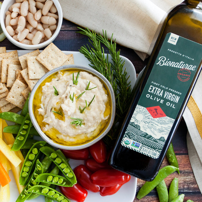 White bean and rosemary spread in a bowl on a platter with crackers, snap peas, cherry tomatoes, and a bottle of olive oil