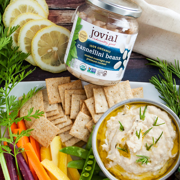 White bean rosemary spread in a bowl with olive oil, surrounded by crackers, vegetables, and a jar of Jovial cannellini beans