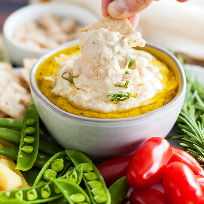 Close-up of white bean rosemary spread in a bowl, creamy texture with olive oil and rosemary garnish, crackers and snap peas nearby